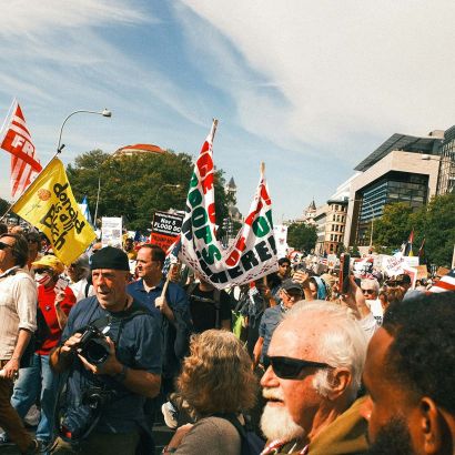 Large crowd at an outdoor protest holding American flags and signs, including "Fake Kings," "I Checked the Constitution — Still No King," and a "Flood DC" banner, with government buildings in the background.